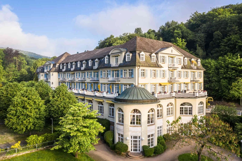 Au&szlig;enansicht der Oberberg Parkklinik Wiesbaden-Schlangenbad: Denkmalgesch&uuml;tztes Geb&auml;ude vor gr&uuml;nen B&auml;umen und blauem Himmel, mit Gehweg im Vordergrund